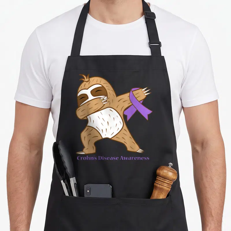 Crohn ribbon apron - Cartoon brown sloth dabbing holding purple ribbon with Crohn's Disease Awareness text — shown on a black cotton canvas apron against a white background