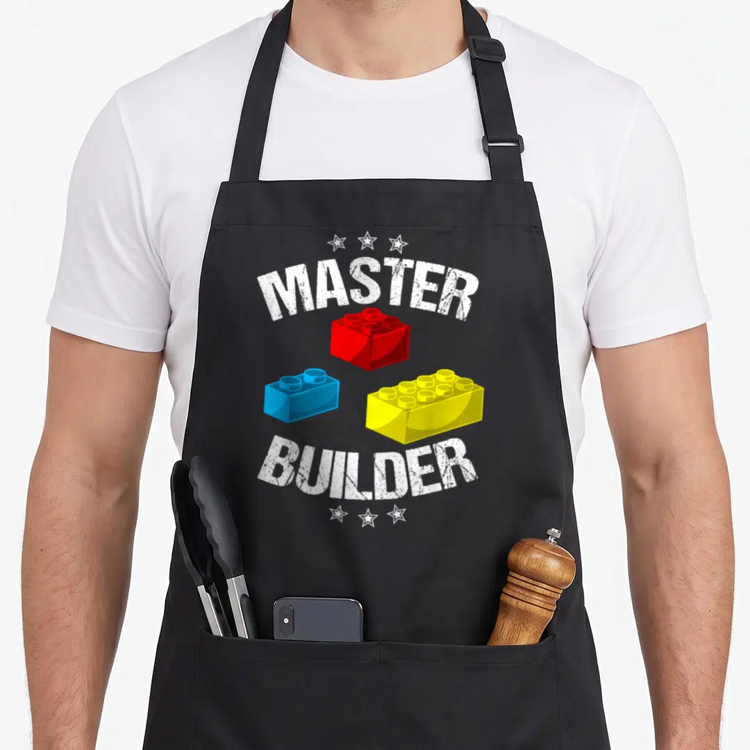 Builder grill master apron - Arched MASTER BUILDER text above red blue yellow building blocks and six white stars — shown on a black cotton canvas apron against a white background