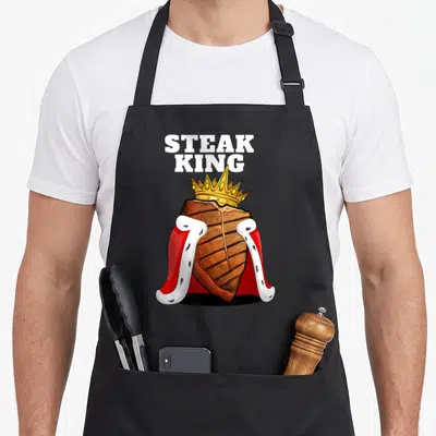 Steak king apron - Cartoon brown steak wearing gold crown and red cape with white trim below white text — shown on a black cotton canvas apron against a white background