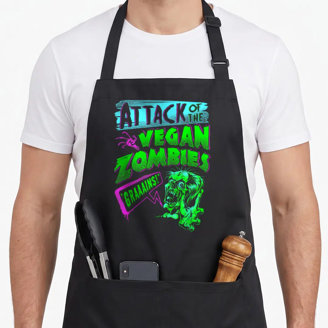 Attack vegan apron - Stacked ATTACK OF THE VEGAN ZOMBIES text with pink spider and green zombie head in cartoon style — shown on a black cotton canvas apron against a white background