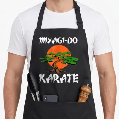 Miyagi tree apron - Cartoon bonsai tree on orange sun with white MIYAGI-DO and KARATE text — shown on a black cotton canvas apron against a white background