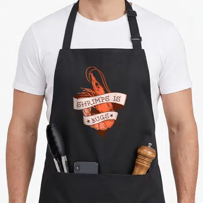 Shrimp meme apron - Cartoon orange shrimp wrapped in ribbon reading SHRIMPS IS BUGS with scattered stars — shown on a black cotton canvas apron against a white background