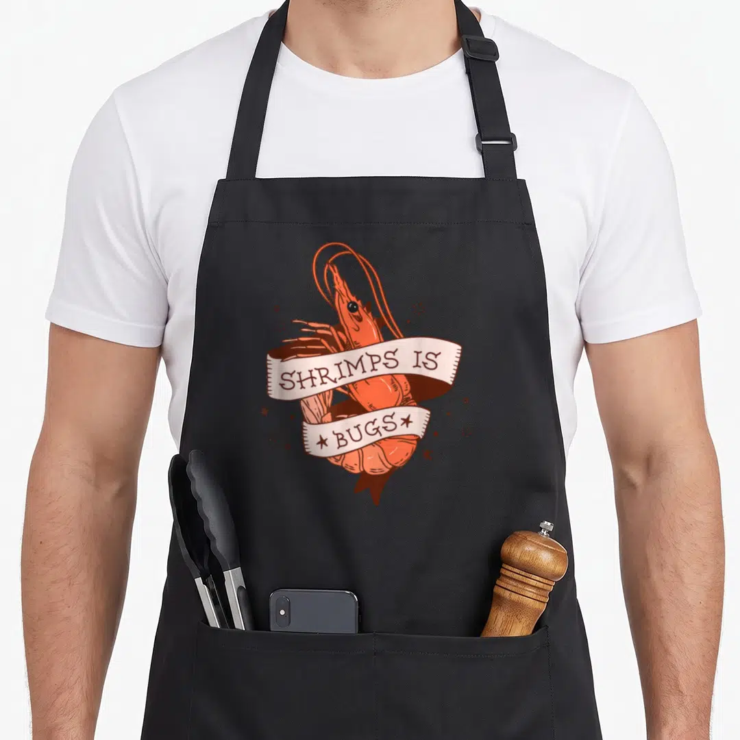 Shrimp meme apron - Cartoon orange shrimp wrapped in ribbon reading SHRIMPS IS BUGS with scattered stars — shown on a black cotton canvas apron against a white background