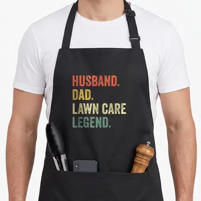 Care lawn apron - Stacked text HUSBAND DAD LAWN CARE LEGEND in orange yellow beige green distressed font — shown on a black cotton canvas apron against a white background