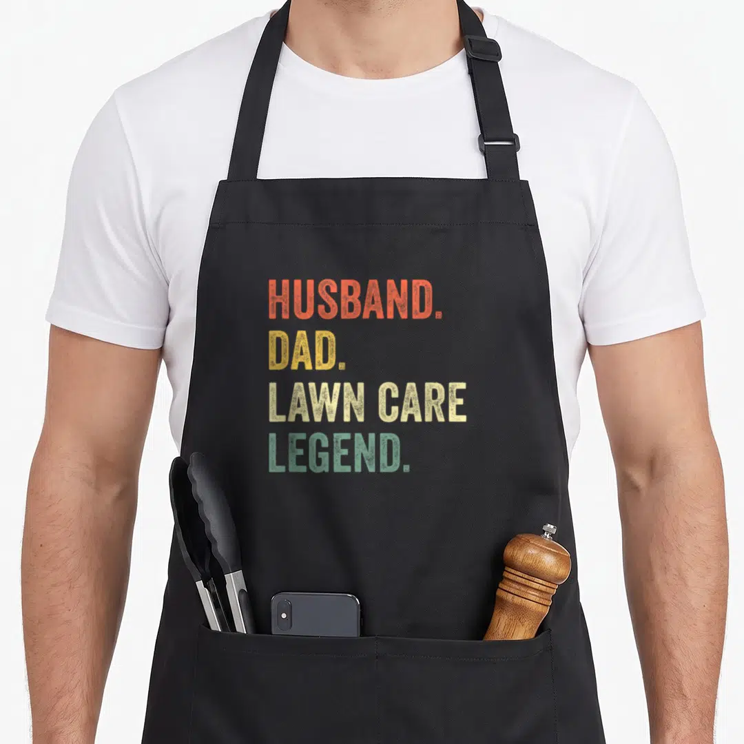 Care lawn apron - Stacked text HUSBAND DAD LAWN CARE LEGEND in orange yellow beige green distressed font — shown on a black cotton canvas apron against a white background