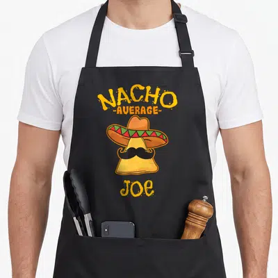Average taco apron - Nacho chip with sombrero mustache and text NACHO AVERAGE JOE in yellow orange — shown on a black cotton canvas apron against a white background