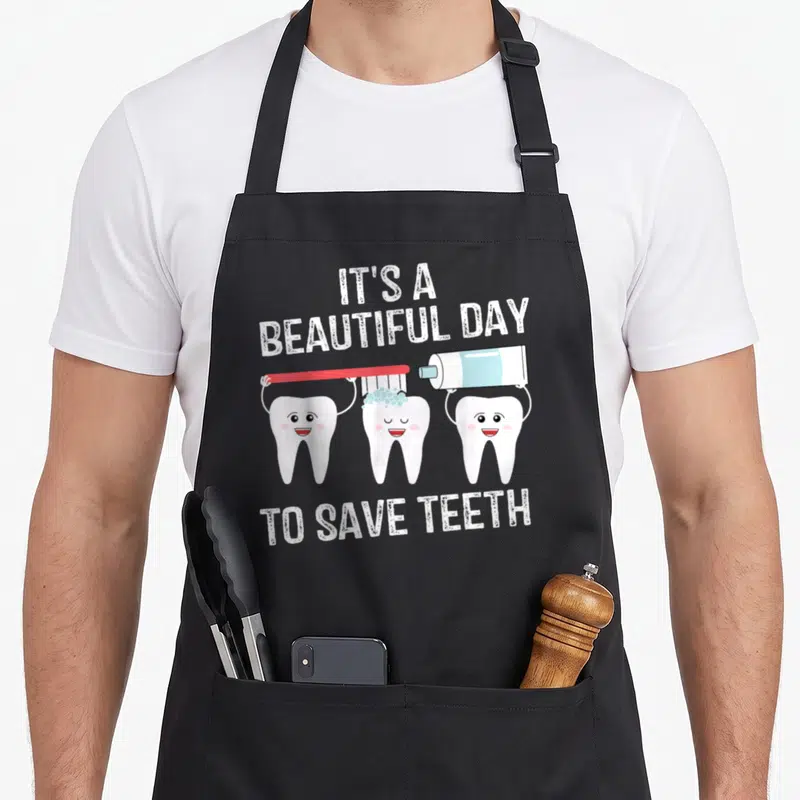 Beautiful dentist apron - Three cartoon teeth brushing and applying paste under the text It's a Beautiful Day to Save — shown on a black cotton canvas apron against a white background