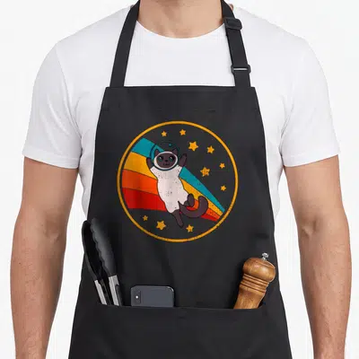 Bicolor cute cat aprons - Retro Siamese cat in space helmet floating over orange circle with colored stripes and stars — shown on a black cotton canvas apron against a white background