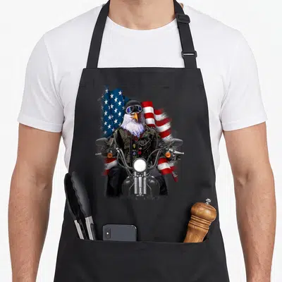 Biker eagles apron - Realistic eagle in biker jacket riding motorcycle before waving American flag in red white blue — shown on a black cotton canvas apron against a white background