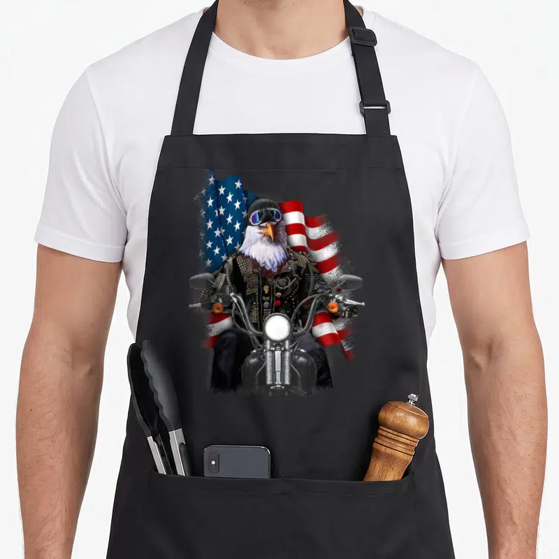 Biker eagles apron - Realistic eagle in biker jacket riding motorcycle before waving American flag in red white blue — shown on a black cotton canvas apron against a white background