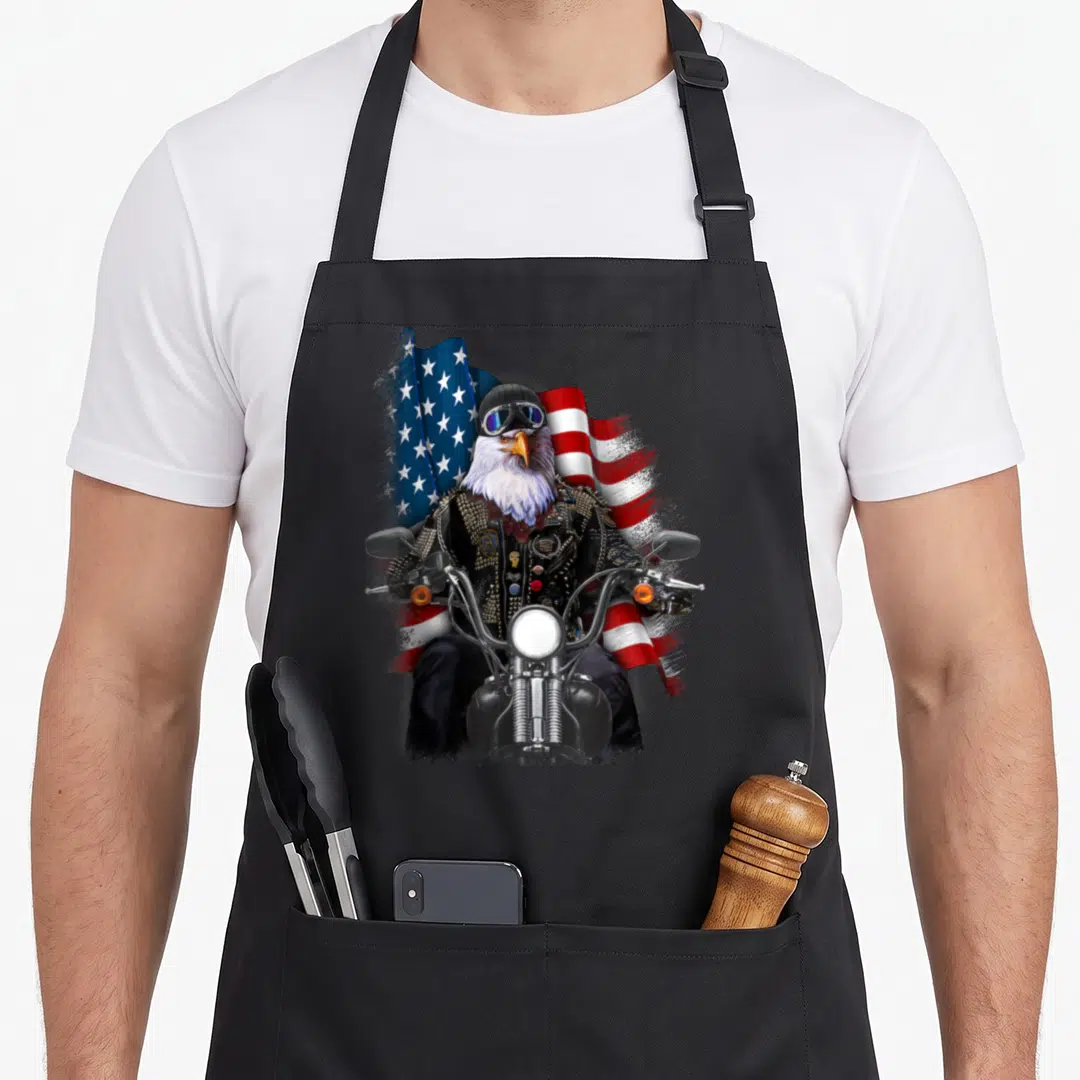 Biker eagles apron - Realistic eagle in biker jacket riding motorcycle before waving American flag in red white blue — shown on a black cotton canvas apron against a white background