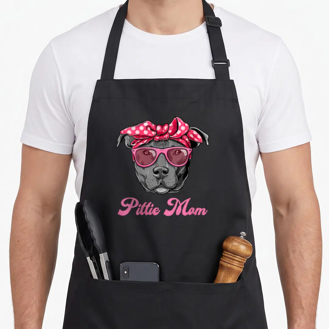 Pittie pitbull dog mom apron - Cartoon grey pit bull head with pink sunglasses and polka dot bow above pink script text — shown on a black cotton canvas apron against a white background