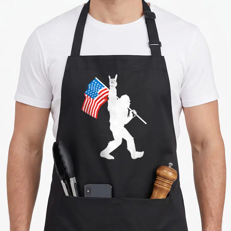 Roll bigfoot apron - White Bigfoot silhouette holding US flag and making rock-on sign — shown on a black cotton canvas apron against a white background