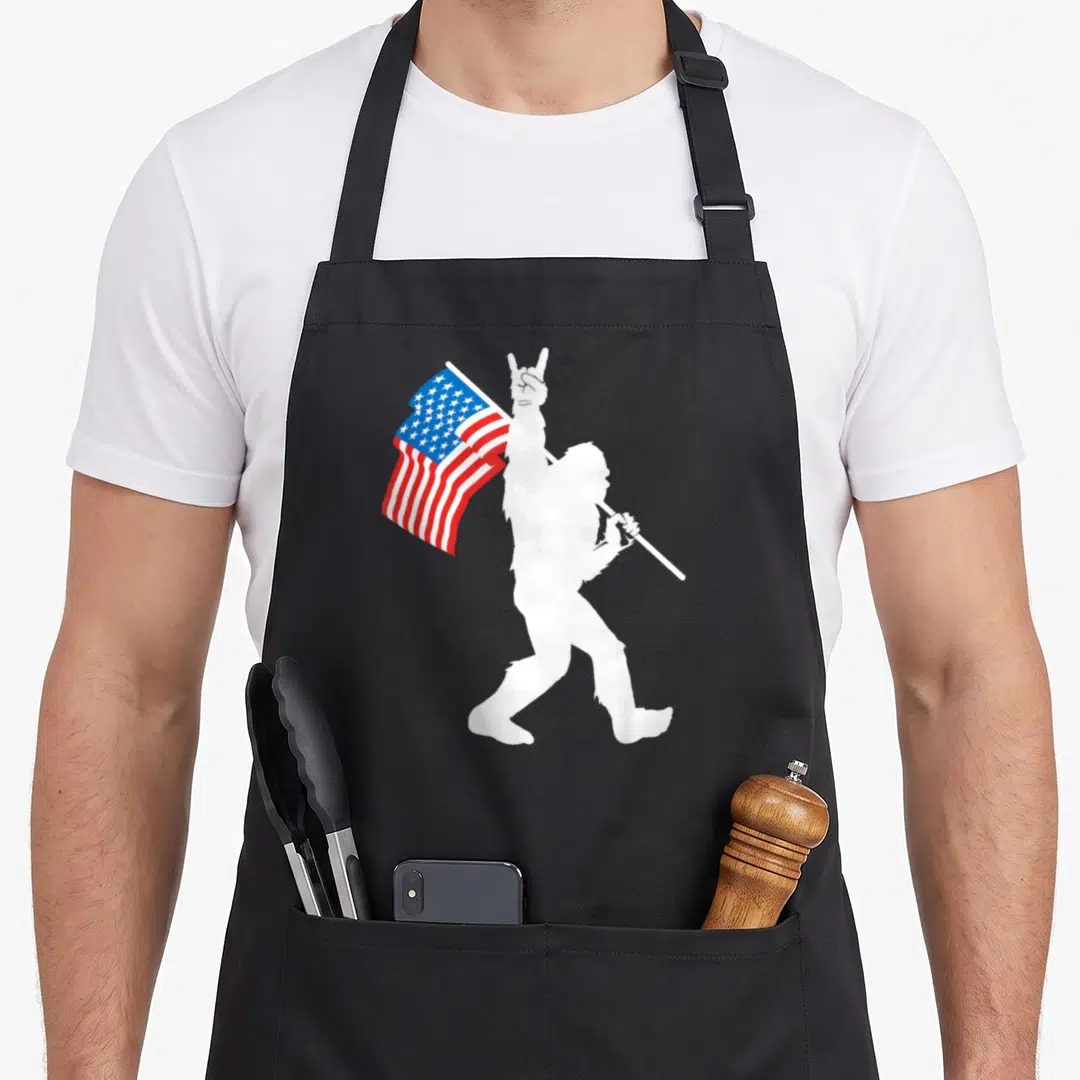 Roll bigfoot apron - White Bigfoot silhouette holding US flag and making rock-on sign — shown on a black cotton canvas apron against a white background
