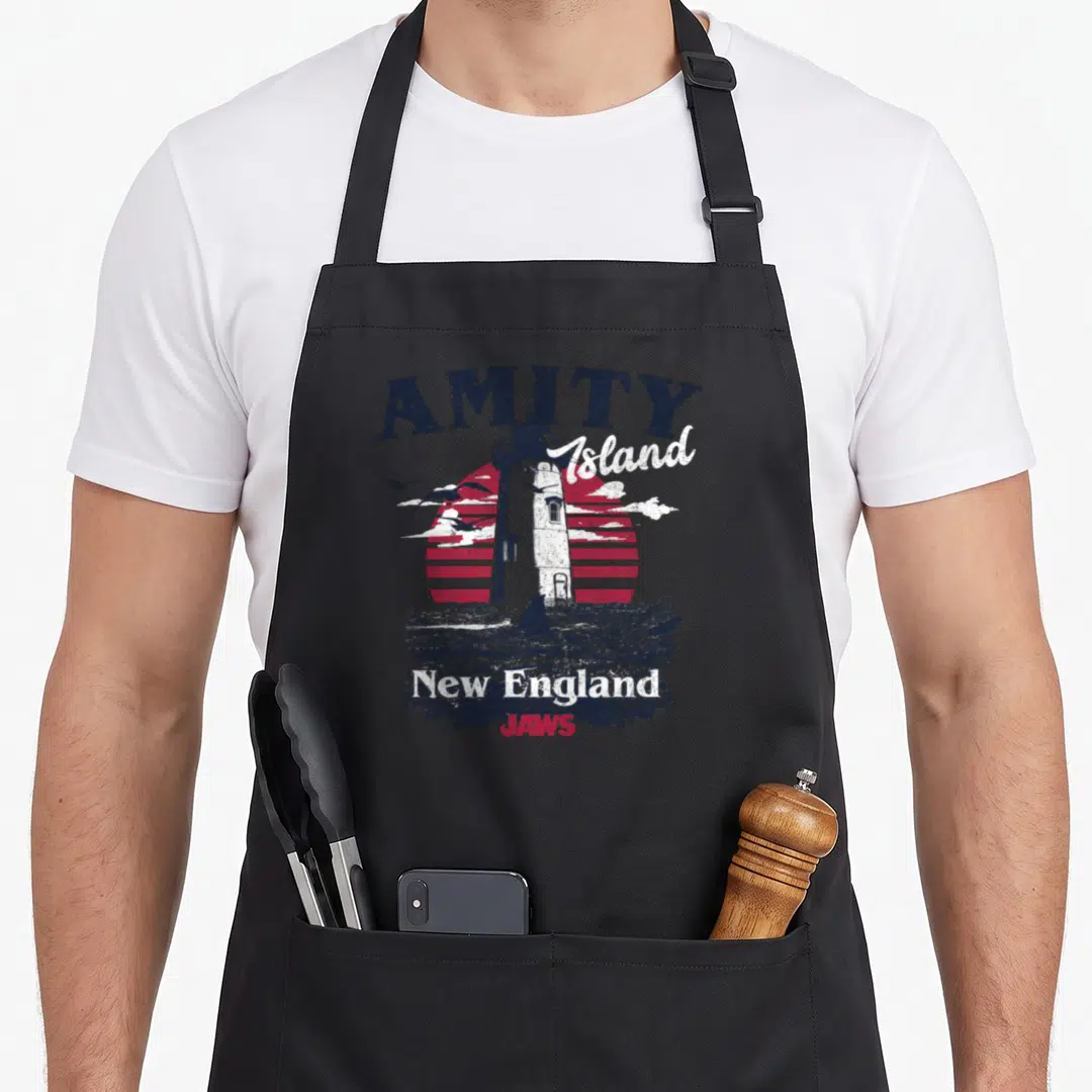 Island england apron - Distressed lighthouse with red striped sun, shark fin, and Amity Island New England JAWS text — shown on a black cotton canvas apron against a white background
