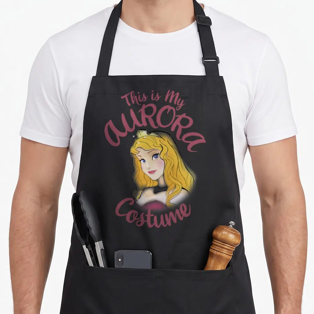 Sleeping halloween apron - Aurora cartoon portrait with This is My AURORA Costume text in pink and gold — shown on a black cotton canvas apron against a white background