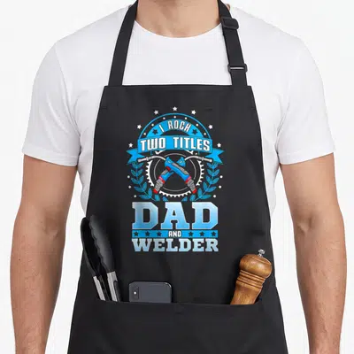 Retro welding welder dad apron - Crossed welding torches over gear with I ROCK TWO TITLES DAD AND WELDER text — shown on a black cotton canvas apron against a white background