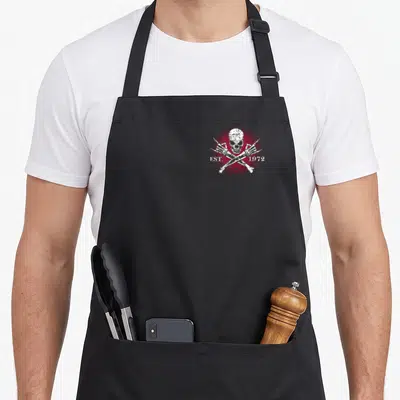 Gen badass apron - White distressed skull and crossbones with EST. 1972 text and red star on dark background — shown on a black cotton canvas apron against a white background