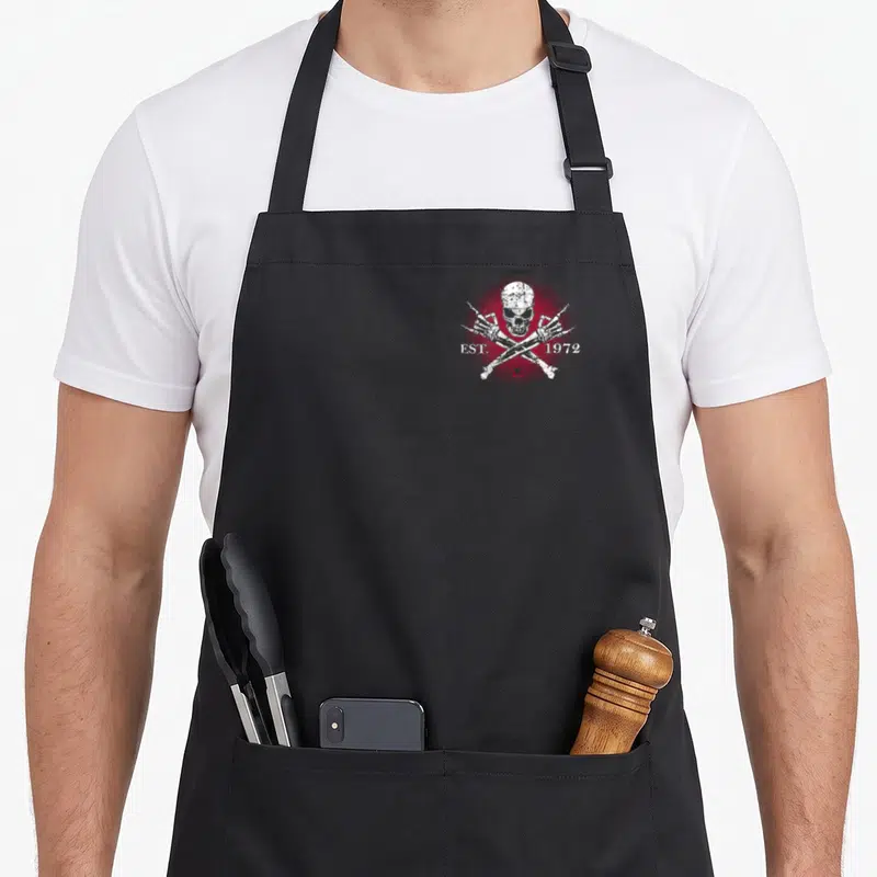Gen badass apron - White distressed skull and crossbones with EST. 1972 text and red star on dark background — shown on a black cotton canvas apron against a white background