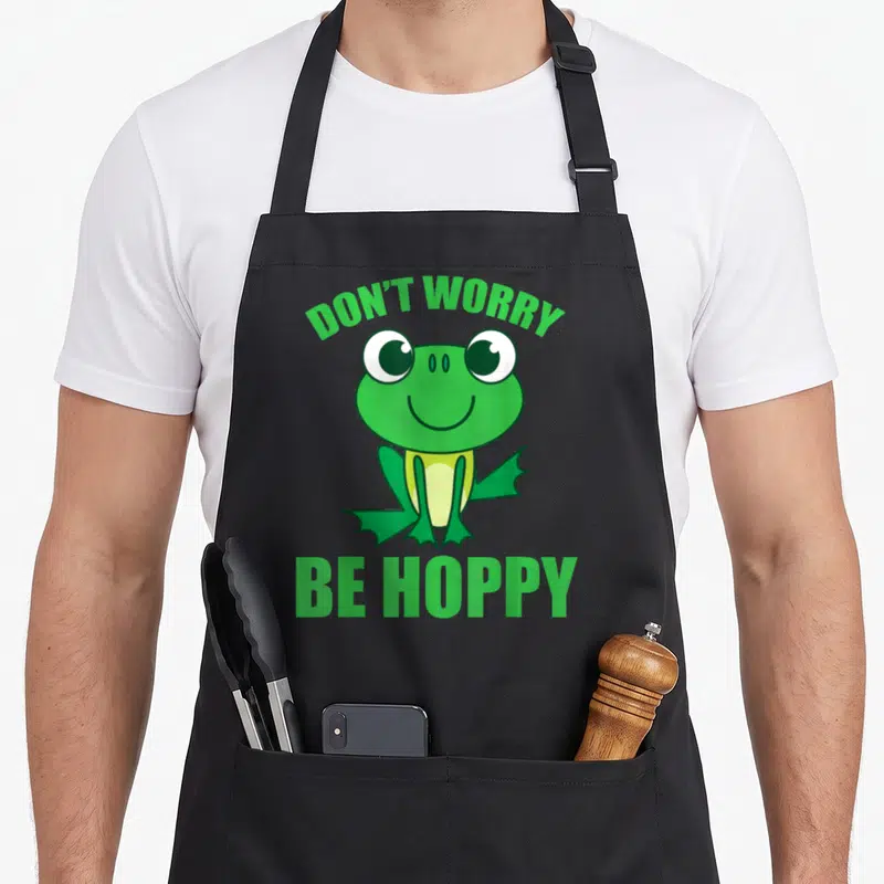 Worry frog aprons - Cartoon frog graphic with green text DON'T WORRY BE HOPPY — shown on a black cotton canvas apron against a white background