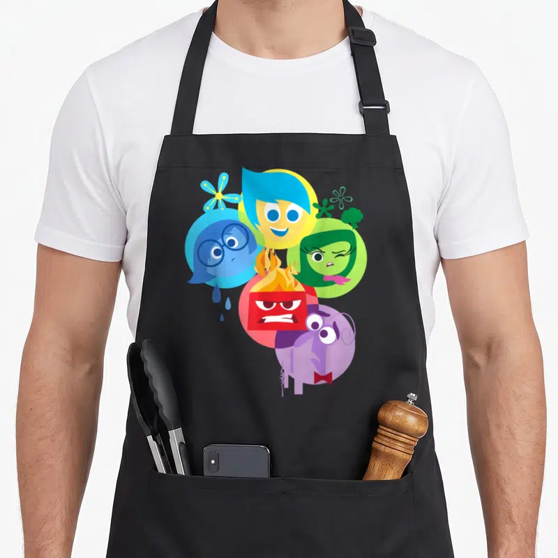 Shot simple apron - Five overlapping cartoon heads in blue yellow green red and purple — shown on a black cotton canvas apron against a white background