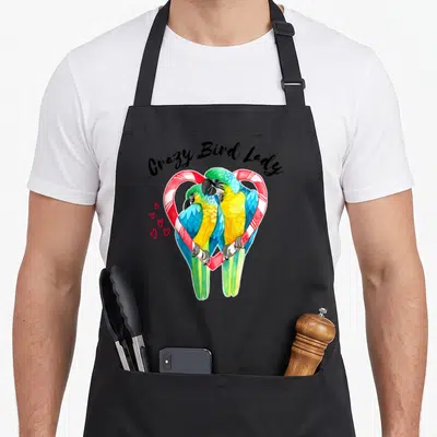 Macaw bird apron - Crazy Bird Lady text above two parrots in red and white striped heart — shown on a black cotton canvas apron against a white background