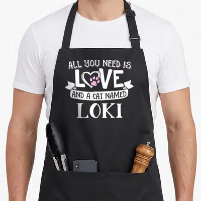 Funny figure mom apron - Stacked white text All You Need Is Love And A Cat Named Loki with pink paw print heart — shown on a black cotton canvas apron against a white background