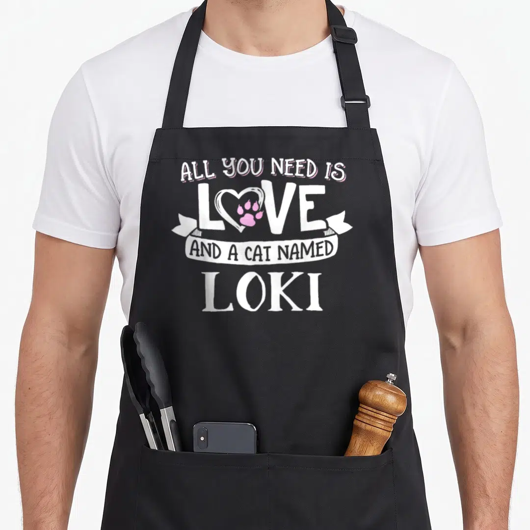 Funny figure mom apron - Stacked white text All You Need Is Love And A Cat Named Loki with pink paw print heart — shown on a black cotton canvas apron against a white background