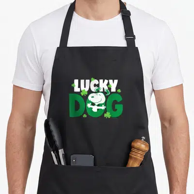 Dog st patrick day aprons - LUCKY DOG text with Snoopy in O and green shamrocks — shown on a black cotton canvas apron against a white background