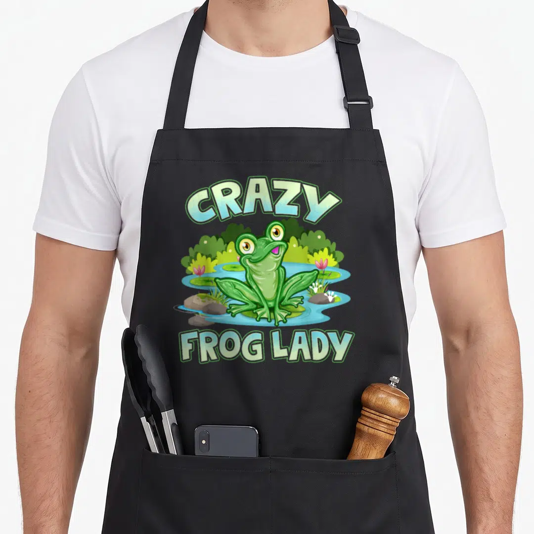 Frog aprons gift - CRAZY FROG LADY text with cartoon frog in pond with lily pads and flowers — shown on a black cotton canvas apron against a white background