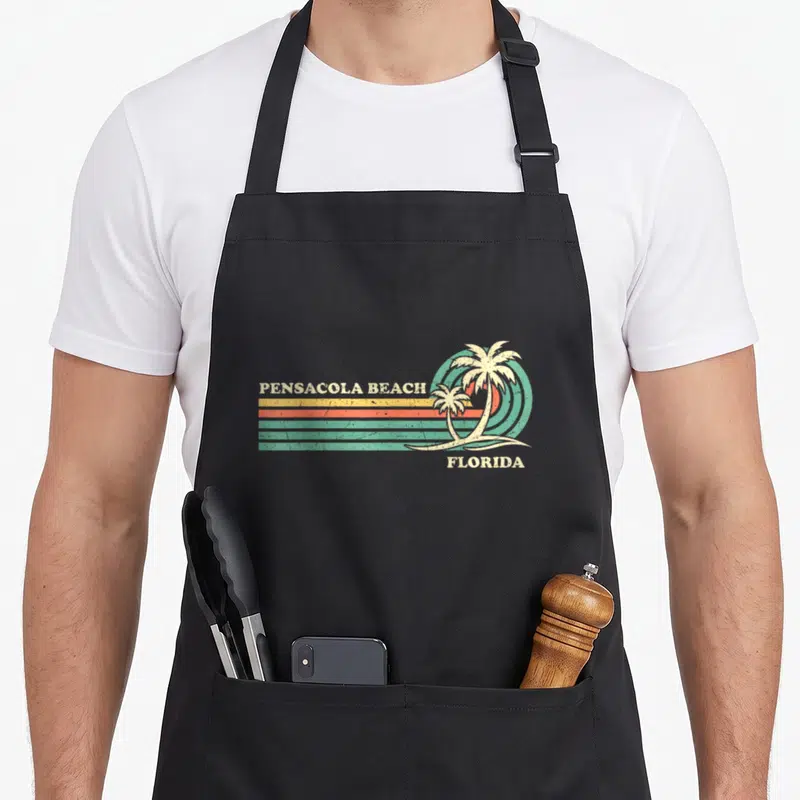 Vacation florida summer aprons - Retro Pensacola Beach Florida print with palm trees and striped sunset in beige orange — shown on a black cotton canvas apron against a white background