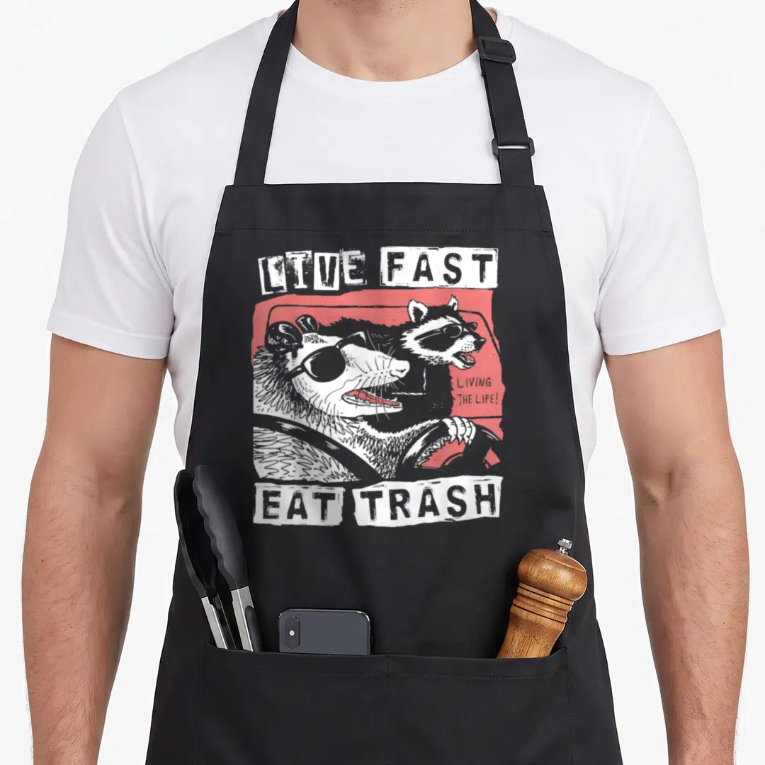 Possum cat apron - Raccoon and possum in red car with live fast eat trash text — shown on a black cotton canvas apron against a white background