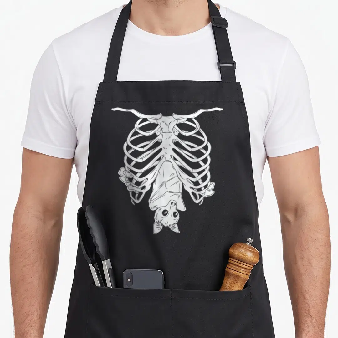 Creepy gothic apron - White line-art ribcage containing an upside-down bat illustration on grey background — shown on a black cotton canvas apron against a white background