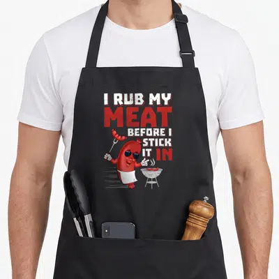 Rub meat aprons - Cartoon red sausage wearing sunglasses and apron holding a fork next to a grill with stacked red — shown on a black cotton canvas apron against a white background