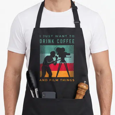 Filmmaker coffee apron - Cinematographer silhouette holding coffee cup against colored stripes with quote text — shown on a black cotton canvas apron against a white background