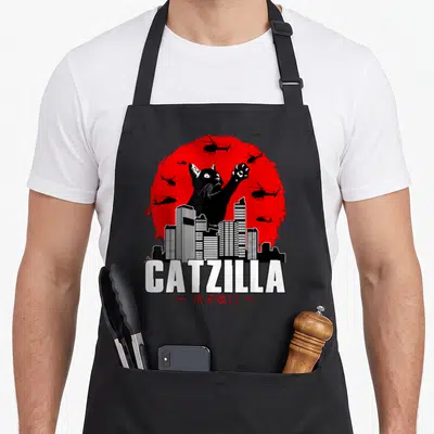 Catzilla cat in an apron - Giant black cat silhouette behind white city buildings and helicopters against a red sun — shown on a black cotton canvas apron against a white background
