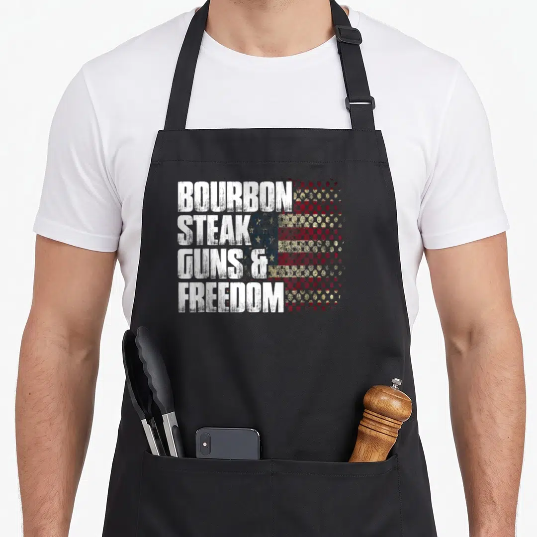 Bourbon gun apron - Stacked white BOURBON STEAK GUNS & FREEDOM text over a red blue and gold halftone USA flag pattern — shown on a black cotton canvas apron against a white background