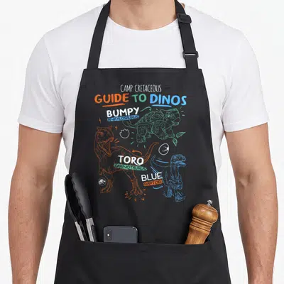 Jurassic line apron - Camp Cretaceous Guide to Dinos print with Bumpy Ankylosaurus, Toro Carnotaurus, and Blue Raptor — shown on a black cotton canvas apron against a white background