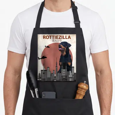 Rottweiler dog apron - Giant rottweiler dog looming over city skyline under red sun with two helicopters flying left — shown on a black cotton canvas apron against a white background