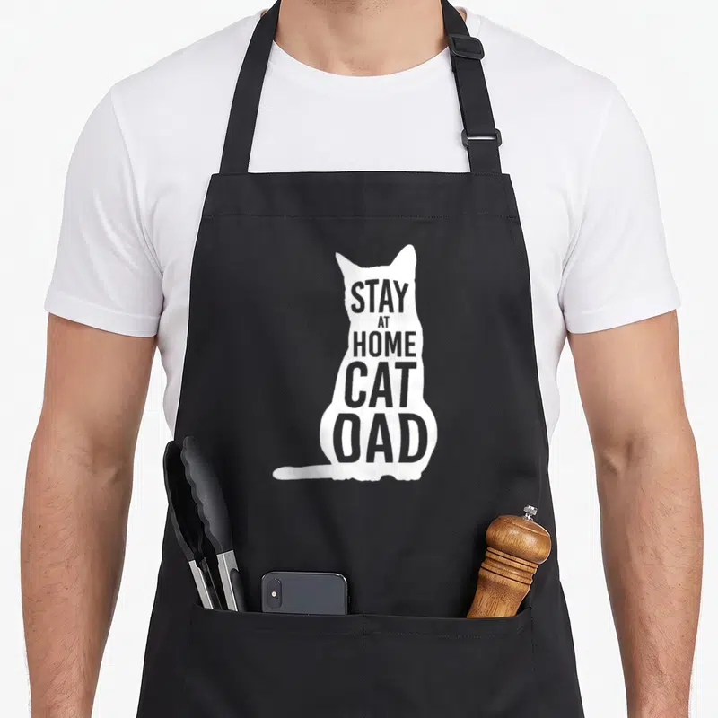 Cat stay at home dad apron - White sitting cat silhouette with grey text STAY AT HOME CAT DAD inside — shown on a black cotton canvas apron against a white background