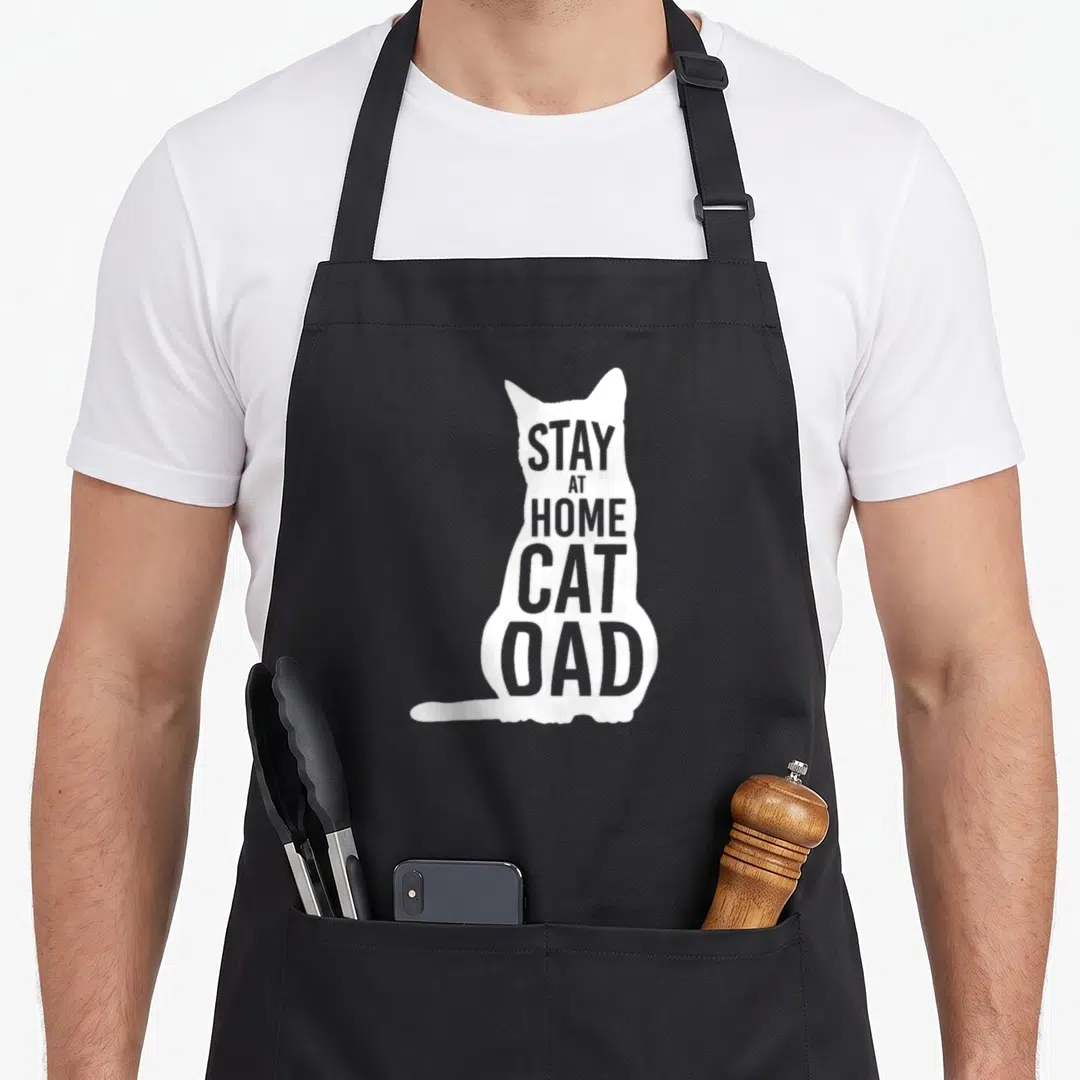Cat stay at home dad apron - White sitting cat silhouette with grey text STAY AT HOME CAT DAD inside — shown on a black cotton canvas apron against a white background
