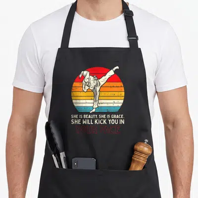 Kick grace apron - Martial artist kicking inside red orange yellow cream blue striped circle with sarcastic text below — shown on a black cotton canvas apron against a white background