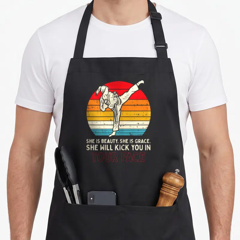 Kick grace apron - Martial artist kicking inside red orange yellow cream blue striped circle with sarcastic text below — shown on a black cotton canvas apron against a white background