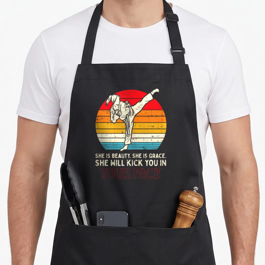 Kick grace apron - Martial artist kicking inside red orange yellow cream blue striped circle with sarcastic text below — shown on a black cotton canvas apron against a white background