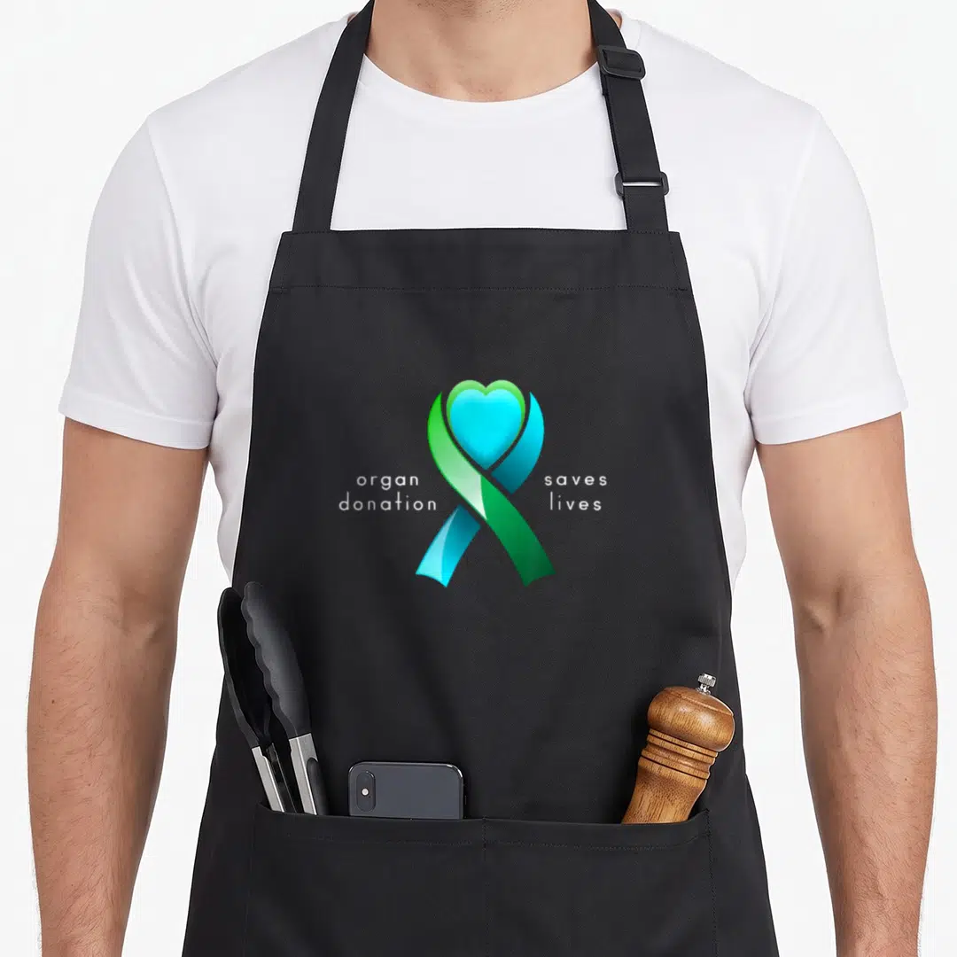 Live organ apron - Green blue ribbon with blue heart and white text organ donation saves lives — shown on a black cotton canvas apron against a white background