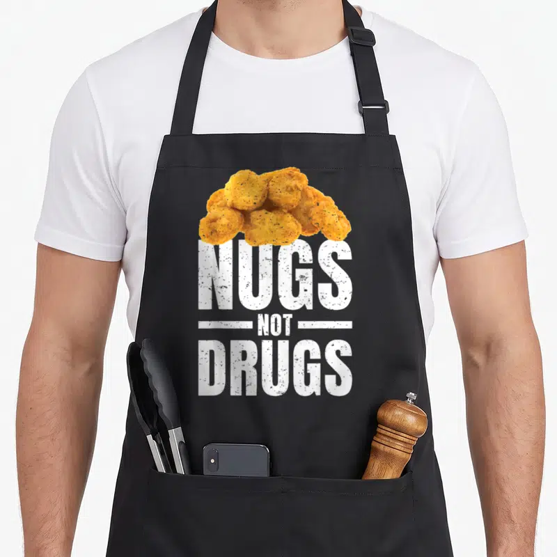Nug chicken print apron - Pile of golden brown chicken nuggets above white NUGS NOT DRUGS text — shown on a black cotton canvas apron against a white background