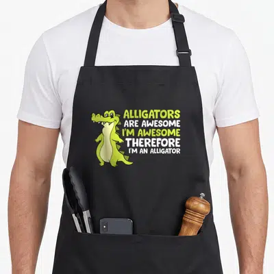 Cute alligator apron - Lime green cartoon alligator standing next to stacked white and lime text reading Alligators Are — shown on a black cotton canvas apron against a white background