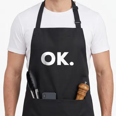 Funny okroshka apron - White OK period text on dark background — shown on a black cotton canvas apron against a white background