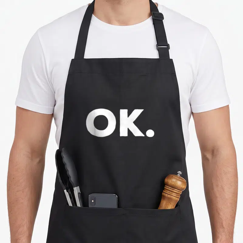 Funny okroshka apron - White OK period text on dark background — shown on a black cotton canvas apron against a white background