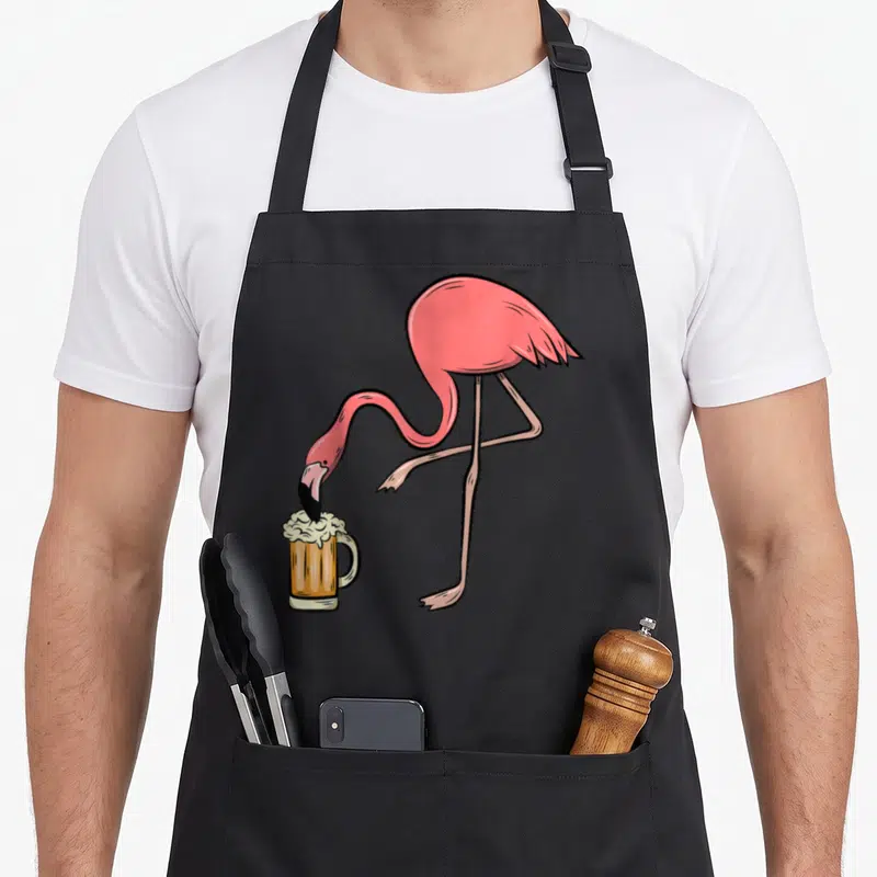 Drinking pink flamingo apron - Pink cartoon flamingo standing on one leg drinking from a foamy beer mug — shown on a black cotton canvas apron against a white background